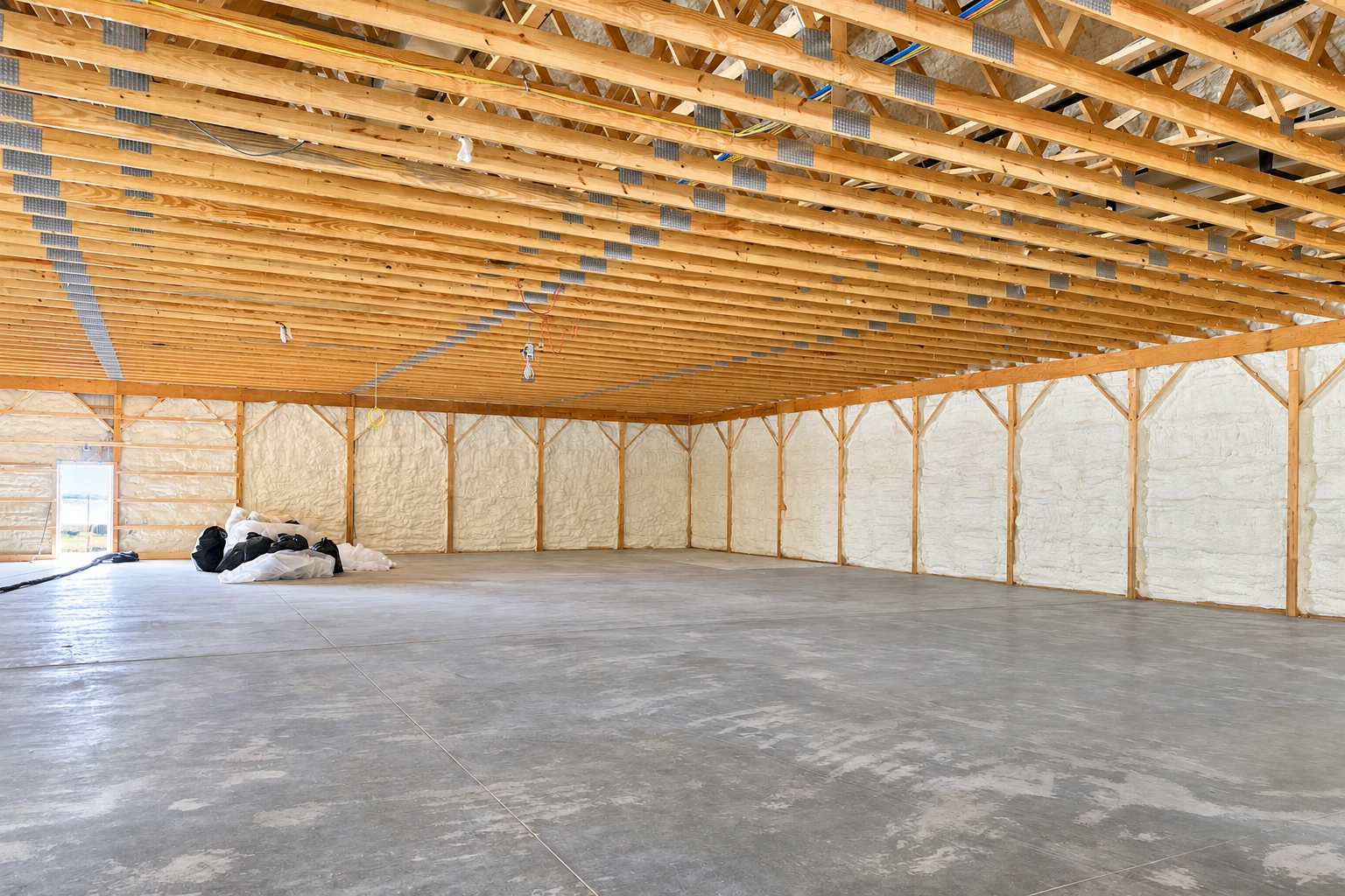 Closed-cell spray foam coats the walls of a large unfinished building beneath exposed wooden roof trusses.