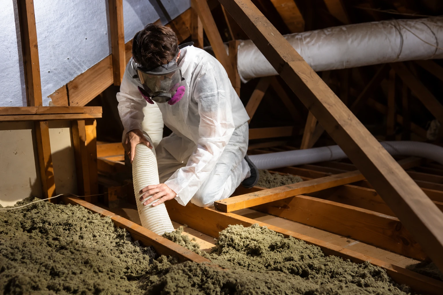 A worker in protective coveralls and a respirator removes attic insulation using a flexible hose.