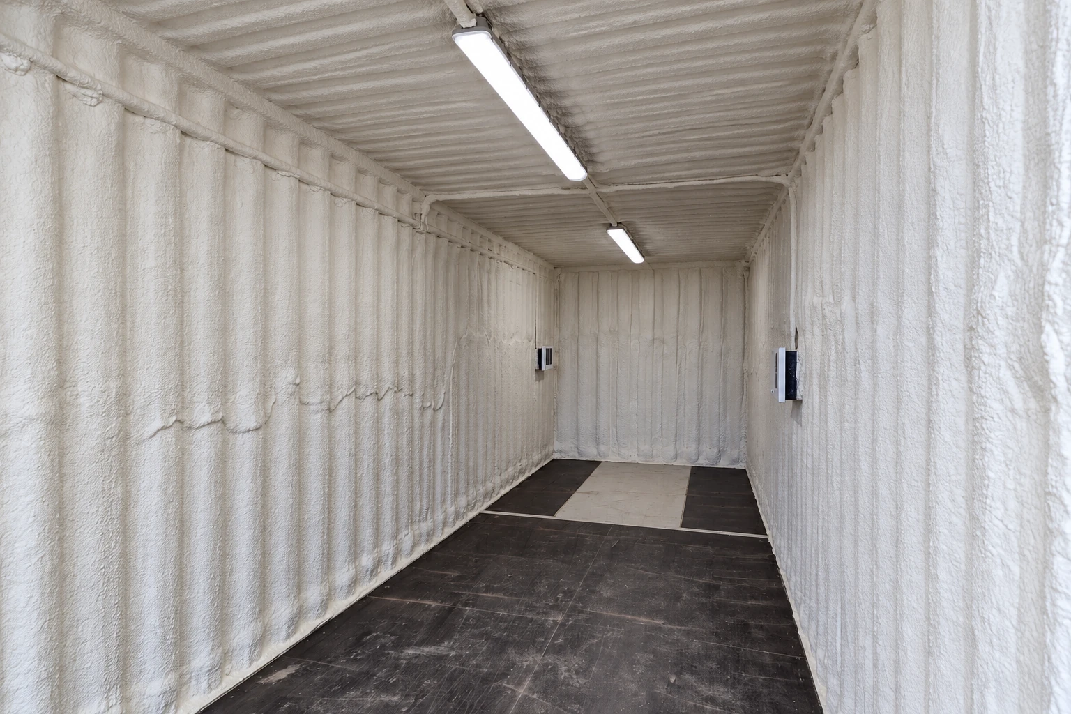 Spray foam coats the walls and ceiling of a narrow room with a dark floor and two overhead lights.