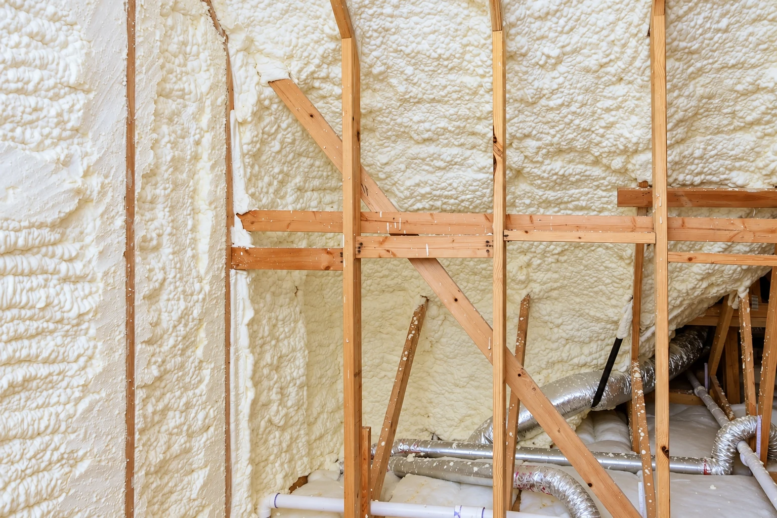 White spray foam coats the interior wall cavities around exposed wooden framing, with ducts and pipes below.